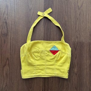 May include: A bright yellow halter top featuring a diamond-shaped patch on the upper right side, divided into a light blue top half and a red bottom half. The garment has a button-fastened neck strap and visible vertical seams across the front.