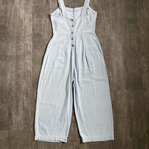 May include: Pale blue denim jumpsuit with wide legs and a button-up front. The jumpsuit has adjustable straps and a fitted bodice. The garment is made of a lightweight fabric, suitable for warmer weather.