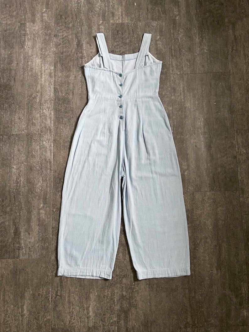 May include: Pale blue denim jumpsuit with wide legs and a button-up front. The jumpsuit has adjustable straps and a fitted bodice. The garment is made of a lightweight fabric, suitable for warmer weather.