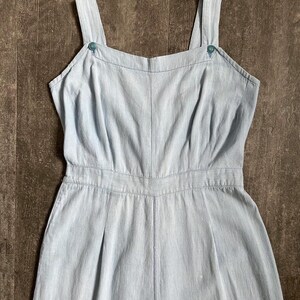 May include: Pale blue jumpsuit with a square neckline and buttoned shoulder straps. The one-piece garment has a fitted waist and wide legs. The fabric has a subtle texture, and the design is simple and classic.