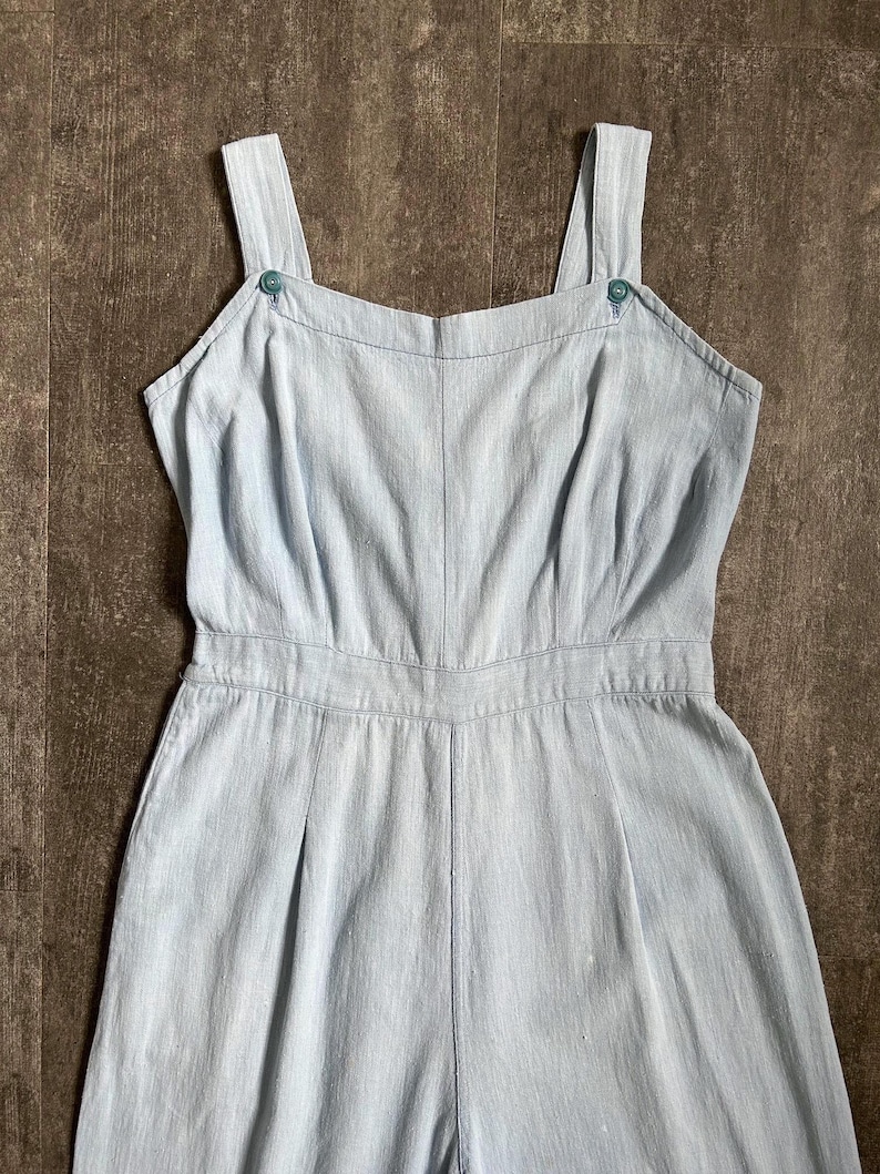 May include: Pale blue jumpsuit with a square neckline and buttoned shoulder straps. The one-piece garment has a fitted waist and wide legs. The fabric has a subtle texture, and the design is simple and classic.