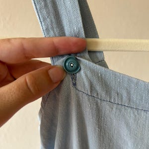 May include: Close-up of a light blue denim garment with a teal snap closure. The garment is hanging on a white hanger. The fabric has a textured appearance and is stitched with blue thread.