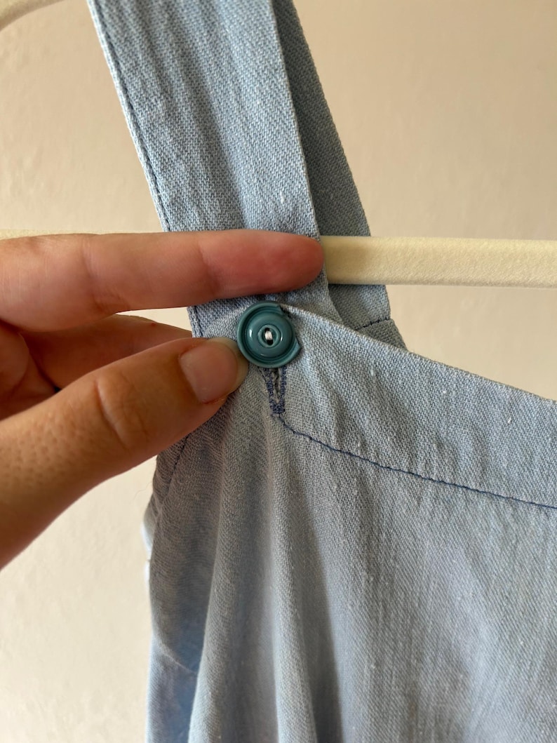 May include: Close-up of a light blue denim garment with a teal snap closure. The garment is hanging on a white hanger. The fabric has a textured appearance and is stitched with blue thread.