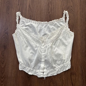 May include: A white vintage camisole with delicate lace trim along the neckline and straps. The camisole features a button-down front and a slightly gathered hem. The fabric appears to be lightweight and semi-sheer, suggesting a vintage or antique style.