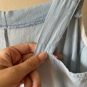 May include: Close-up of a light blue denim garment, possibly a dress or jumpsuit, with visible stitching and shoulder straps. The fabric appears to be a light wash denim. The image highlights the texture and construction of the garment.