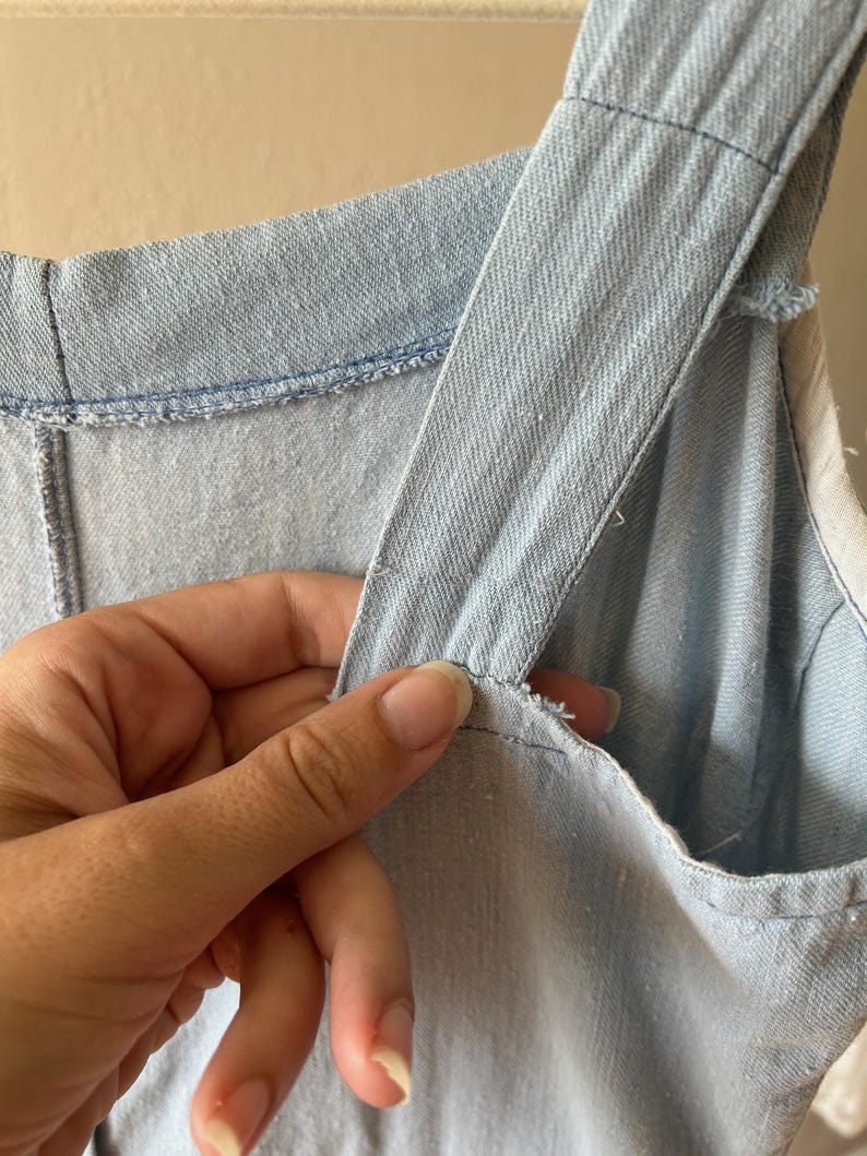 May include: Close-up of a light blue denim garment, possibly a dress or jumpsuit, with visible stitching and shoulder straps. The fabric appears to be a light wash denim. The image highlights the texture and construction of the garment.