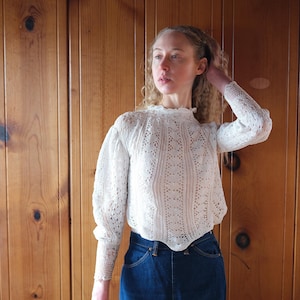 May include: A cream-colored, long-sleeved blouse with a delicate, open-weave pattern. The blouse features a high, ruffled neckline and fitted cuffs. The model is wearing the blouse with dark blue denim jeans. The background is a wooden wall.