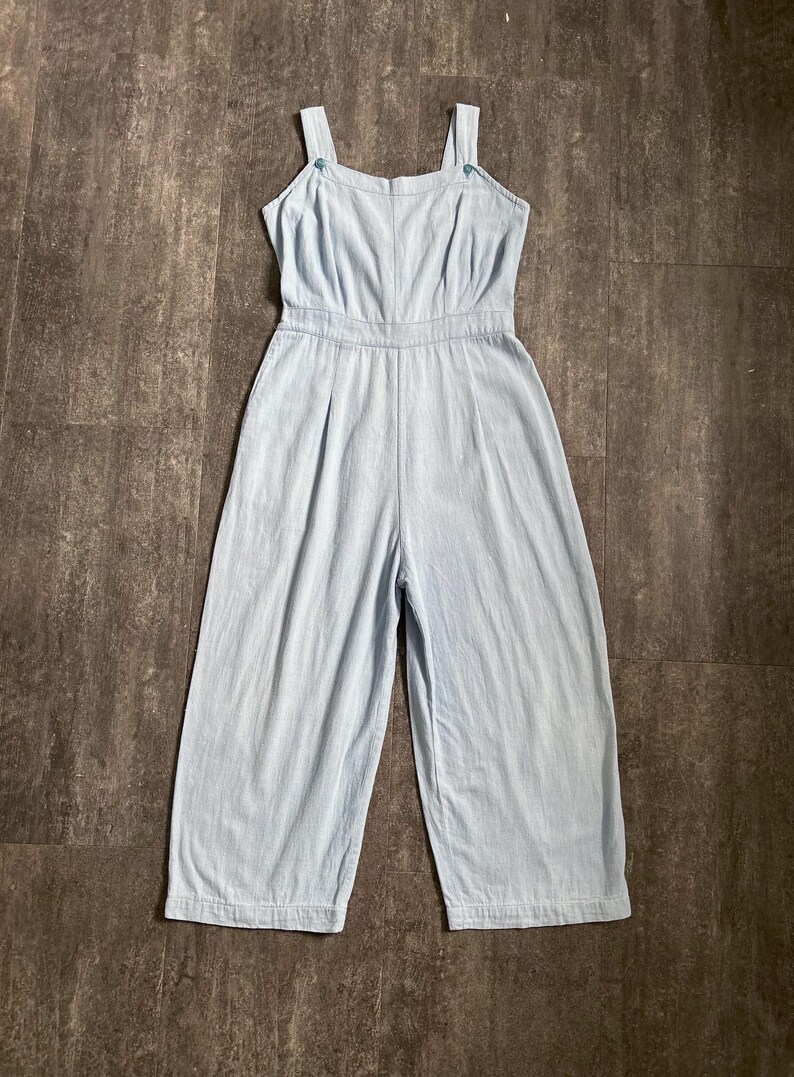 May include: Pale blue jumpsuit with a square neckline and wide legs. The jumpsuit has thin straps with small blue buttons. The waist is fitted, and the fabric looks like a lightweight material, suitable for warmer weather.