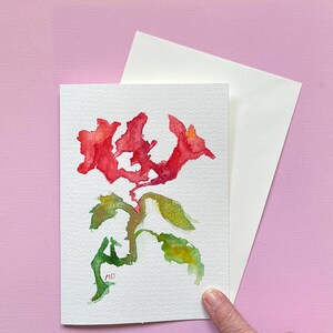 May include: Watercolor greeting card featuring a red floral design with green and yellow leaves. The artwork is on white paper, accompanied by a white envelope. The background is a soft pink. The artist's initials, "MD", are visible on the card.