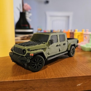 May include: A 3D-printed olive green pickup truck model with black wheels and accents. The toy truck has a detailed front grille, side mirrors, and a textured surface. The model is displayed on a wooden surface.