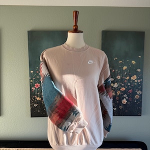 Op de afbeelding: Een lichtroze Nike sweatshirt met mouwen met patronen in blauw, rood en grijs. Het sweatshirt wordt op een mannequin getoond. Een klein wit Nike-logo staat op de linkerbovenborst.