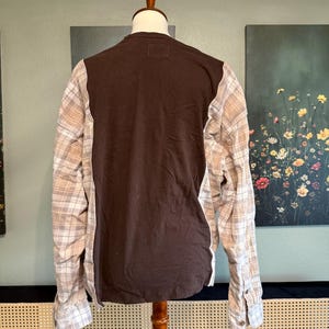 May include: A brown long-sleeved shirt with plaid sleeves. The back of the shirt is solid brown, and the sleeves feature a beige and white plaid pattern. The shirt is displayed on a wooden mannequin.