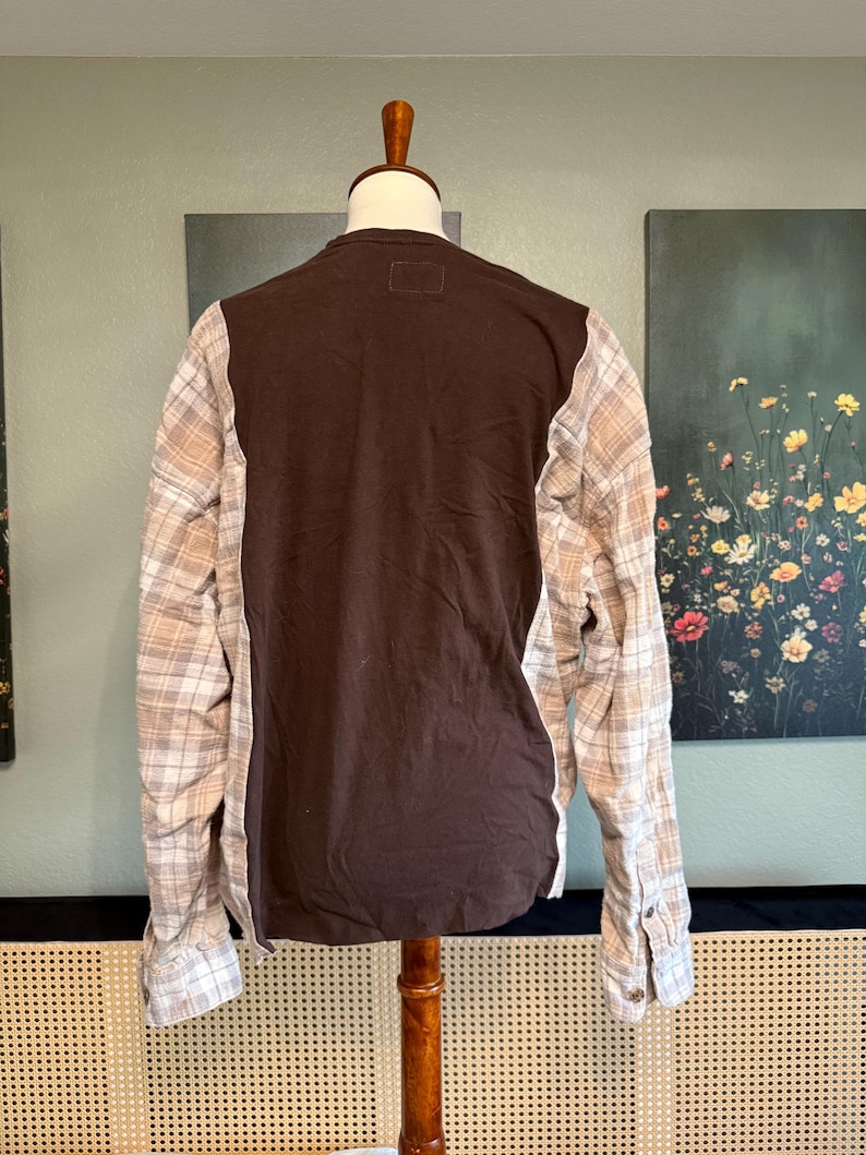 May include: A brown long-sleeved shirt with plaid sleeves. The back of the shirt is solid brown, and the sleeves feature a beige and white plaid pattern. The shirt is displayed on a wooden mannequin.
