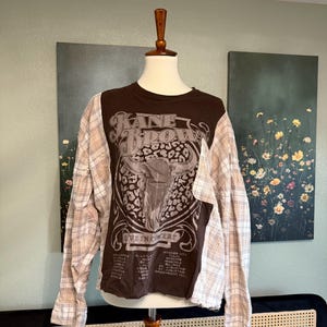 May include: A brown t-shirt with a graphic of a bull's head and the text "KANE BROWN LIVE IN CONCERT". The shirt has long sleeves made from a tan and white plaid fabric.