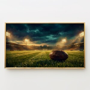 May include: A framed print depicting an American football on a green field in a stadium. The image has a dramatic, artistic style with a dark, cloudy sky and bright stadium lights illuminating the field.