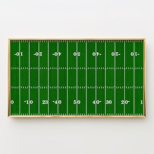 May include: A framed print of a football field, featuring green turf with white yard lines and numbers. The field is marked with yardage indicators from 0 to 50, with a gold-colored frame. A decorative piece for sports enthusiasts.