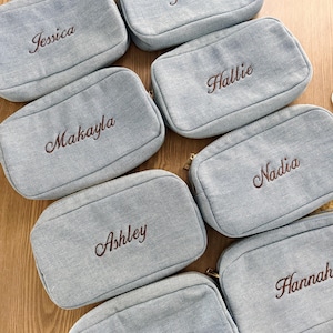 Peut inclure: Plusieurs trousses de maquillage en denim bleu clair, chacune brodée d'un nom différent en écriture marron. Les trousses sont de forme rectangulaire avec une fermeture éclair et une tirette dorée.