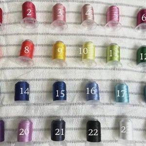 May include: A collection of 24 spools of embroidery thread in various colors, each labeled with a number from 1 to 24. The threads are arranged in rows on a textured, off-white surface.