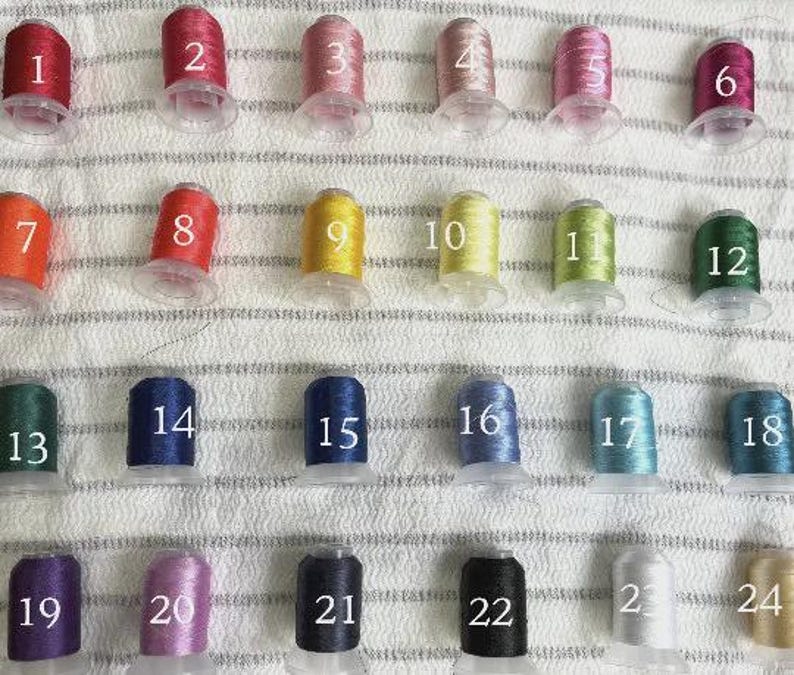 May include: A collection of 24 spools of embroidery thread in various colors, each labeled with a number from 1 to 24. The threads are arranged in rows on a textured, off-white surface.