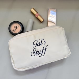 Pode incluir: Uma bolsa de cosméticos creme bordada com "Tal's Stuff" em escrita azul marinho. A bolsa é cercada por itens de maquiagem, incluindo um compacto, batom e um tubo de base.