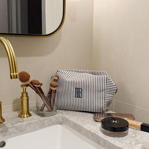 May include: A grey and white striped cosmetic bag with the initials "FHB" embroidered on it sits on a marble countertop. Makeup brushes are in a glass, and other makeup items are visible.