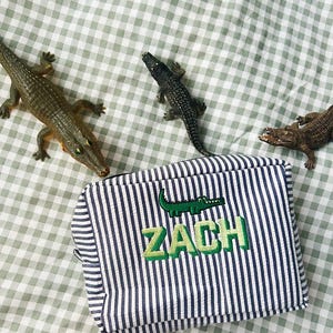 Peut inclure: Une trousse zippée à rayures bleues et blanches avec le nom "ZACH" brodé en vert, ornée d'un appliqué d'alligator vert. Des jouets alligators sont disposés autour de la trousse sur un fond à carreaux verts et blancs.