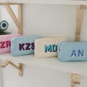 Pode incluir: Quatro bolsas cosméticas retangulares em rosa, azul claro, creme e azul claro, cada uma com um conjunto diferente de letras 3D. As bolsas são exibidas em uma prateleira branca. As letras são ZR, KZS, MD e AN. Uma pequena planta e uma decoração em forma de olho são visíveis.