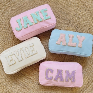 May include: Four rectangular cosmetic bags in pastel shades: pink, pale blue, and cream. Each bag displays a name in glittery, textured letters: "JANE", "ALY", "EVIE", and "CAM". The bags are set on a woven, beige surface.
