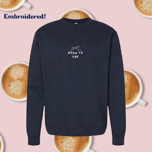 May include: Navy blue sweatshirt with the embroidered text "BORN TO YAP" in pink and a bow design. The word "Embroidered!" is in the top left corner. The background features coffee cups.
