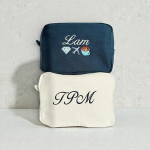 Puede incluir: Dos pequeñas bolsas de viaje rectangulares. La bolsa superior es azul marino con el nombre "Lam" bordado en cursiva, junto con un diamante, un avión y una taza de café. La bolsa inferior es de color blanco roto con "TPM" bordado.