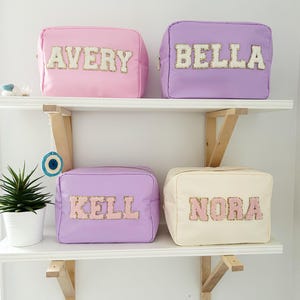 Può includere: Quattro trousse per cosmetici personalizzate in rosa, lilla e crema, ciascuna con un nome scritto con lettere di paillettes. Le borse sono rettangolari con chiusura a zip e sono esposte su una mensola bianca.