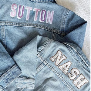 Peut inclure: Deux vestes en jean bleu clair. L'une porte le nom "SUTTON" brodé en violet et blanc. L'autre porte le nom "NASH" brodé en blanc et marron. Les deux vestes ont un design classique de veste en jean.