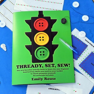 May include: A green book titled "THREADY, SET, SEW!" with a traffic light design made of buttons. The book is surrounded by white buttons, fabric patches, a pen, and a spool of thread, all on a blue surface.