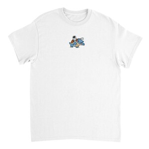 Puede incluir: Camiseta blanca con un pequeño personaje de dibujos animados bordado. El personaje es una criatura azul y marrón con forma de tortuga y vientre amarillo. El diseño está centrado en el pecho de la camiseta.