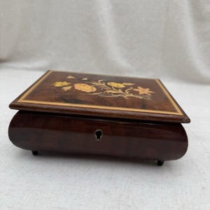 May include: A rectangular wooden music box with a dark brown polished finish and four small feet. The lid features an inlaid floral design in lighter wood tones. A central keyhole is visible on the front panel of the box, which has curved sides.