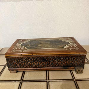 Puede incluir: Una caja rectangular de madera con un panel central de color verde oscuro jaspeado en la tapa, rodeado de incrustaciones de líneas geométricas claras. Los lados están decorados con un patrón de hojas talladas y se apoya sobre pies pequeños.