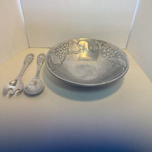 Könnte beinhalten: Eine silberfarbene Servierschüssel mit geprägten Obstmotiven, zusammen mit einer Serviergabel und einem Löffel. Die Schüssel hat eine breite, flache Form. Das Servierbesteck hat dekorative Griffe.