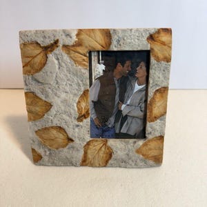 Może przedstawiać: Kwadratowa ramka na zdjęcia z teksturowaną, jasnoszarą powierzchnią, ozdobiona wbudowanymi suszonymi liśćmi. Ramka zawiera zdjęcie pary. Liście są mieszanką brązu i beżu. Ramka ma około 13 cm wysokości i szerokości.