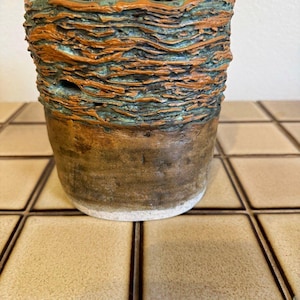 May include: A ceramic planter with a smooth, solid brown base and a textured, uneven upper rim featuring horizontal ridges in alternating shades of copper-orange and deep teal-green. The vessel has a wide, open top and a cylindrical shape.