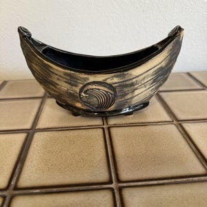 Peut inclure: Un pot en céramique en forme de bateau avec un intérieur noir et un extérieur texturé à la finition dorée et noire vieillie. Le centre du récipient présente un relief circulaire représentant une vague stylisée. La base repose sur des pieds.
