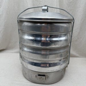 May include: A cylindrical, multi-tiered metal food carrier with a polished silver finish. The container features four stackable compartments, a secure lid, and a sturdy wire handle that locks the sections together for easy transport and storage.