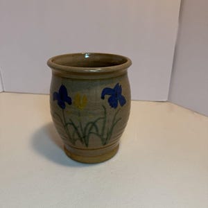 Op de afbeelding: Een handgemaakte keramische vaas met een bruine basis en body. De vaas is versierd met blauwe en gele irissen en groene stelen. De vaas heeft een cilindrische vorm en een licht uitlopende rand. De vaas is geschikt voor het vasthouden van bloemen of als decoratief item.