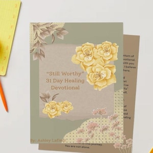 May include: A devotional book titled "Still Worthy" with floral illustrations in yellow and brown tones. The book has a light green background with text and floral accents. An orange pen and yellow notepad are in the upper left corner.