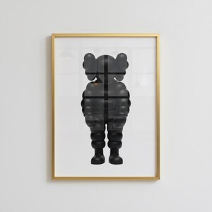 May include: Framed artwork featuring a black, stylized figure with rounded features and a cross-shaped design overlay. The artwork is encased in a gold-colored frame, creating a modern aesthetic. The figure's form is reminiscent of a cartoon character.