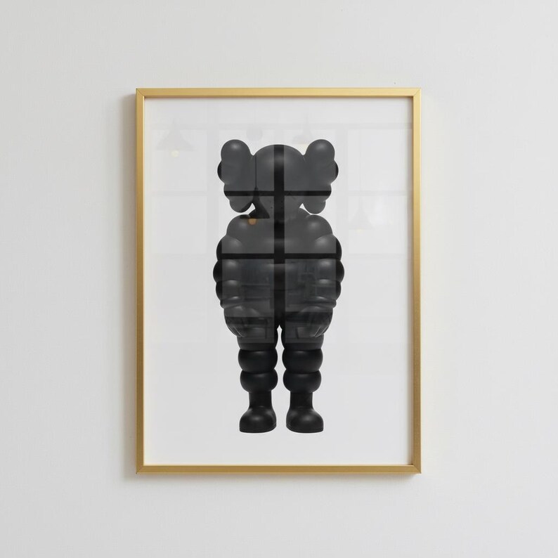 May include: Framed artwork featuring a black, stylized figure with rounded features and a cross-shaped design overlay. The artwork is encased in a gold-colored frame, creating a modern aesthetic. The figure's form is reminiscent of a cartoon character.