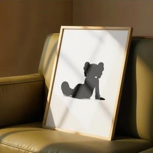 May include: A framed art print featuring a black silhouette of a seated cartoon figure with large, round ears. The artwork is set against a white background and framed in a light wood frame. The print is displayed on a green sofa.