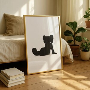 May include: A framed art print featuring a black silhouette of a seated figure with distinctive ears. The artwork is set against a white background within a gold frame. The print leans against a wall on a wooden floor.