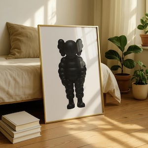 May include: A framed art print featuring a black, stylized figure with large, rounded ears and a puffed-up body. The artwork is set against a white background within a gold frame, leaning against a bed frame and hardwood floor.