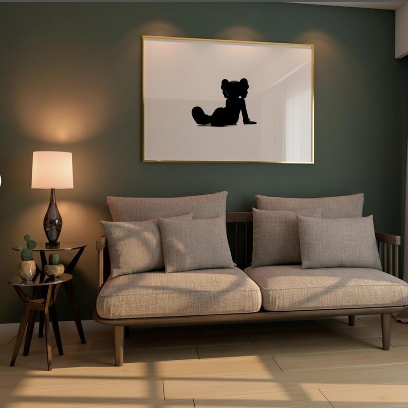 May include: A living room scene featuring a light-colored sofa with several matching pillows. A framed artwork with a black silhouette hangs above the sofa. A small side table with a lamp and decorative plants sits to the left.