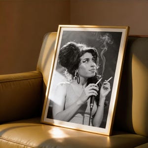 May include: A framed black and white photograph of a singer holding a microphone and a cigarette. The photograph is in a gold frame and is leaning against a leather sofa. The singer is wearing a sleeveless dress and large earrings.